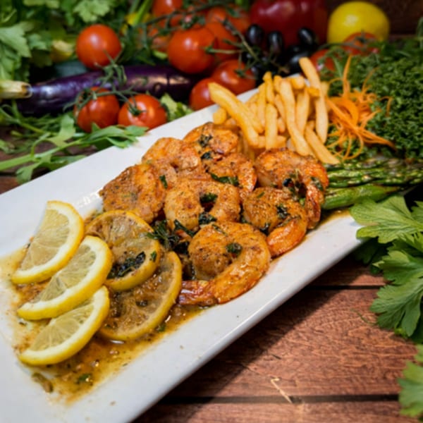 A white platter of grilled shrimp with lemon slices, fries, and asparagus, surrounded by fresh vegetables.